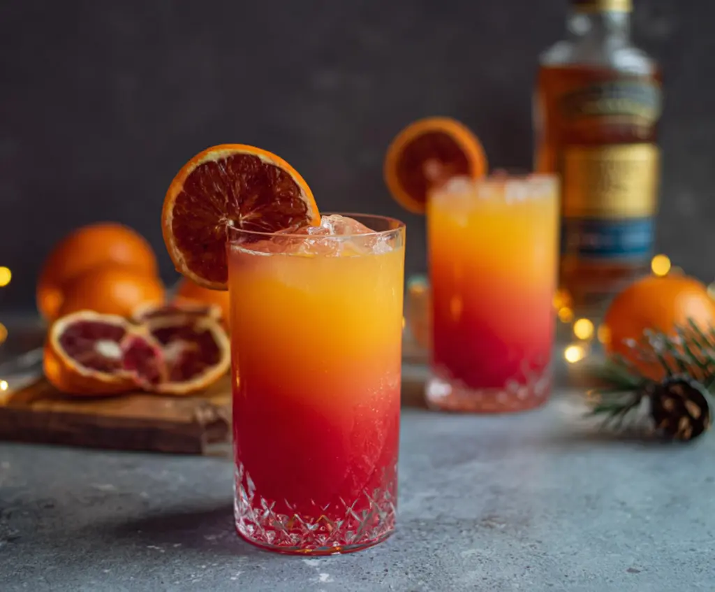 Festive Christmas Tequila Sunrise cocktail with vibrant red and orange layers garnished with cherry and orange slice.