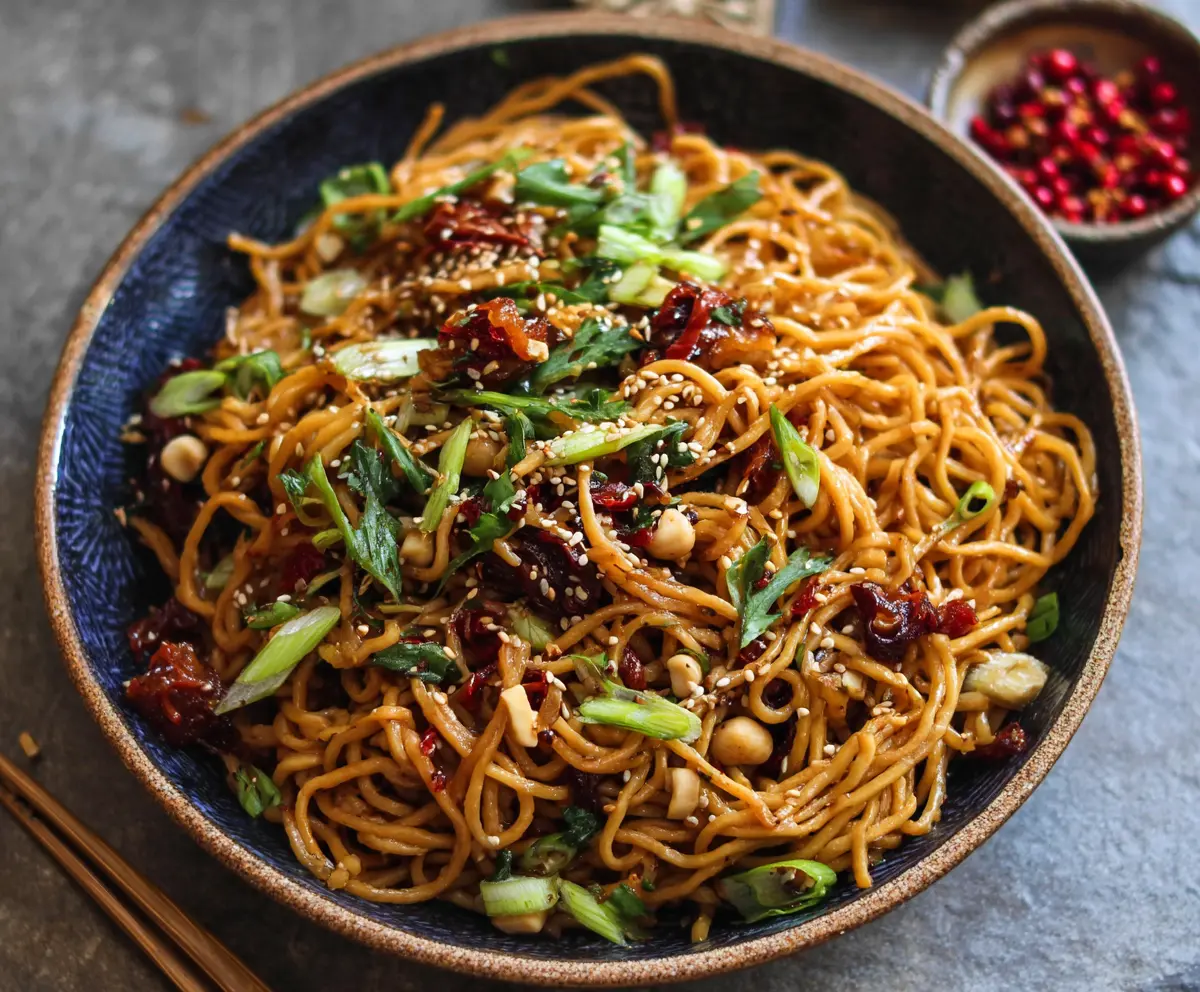 Delicious spicy sesame noodles garnished with green onions and sesame seeds on a white plate.